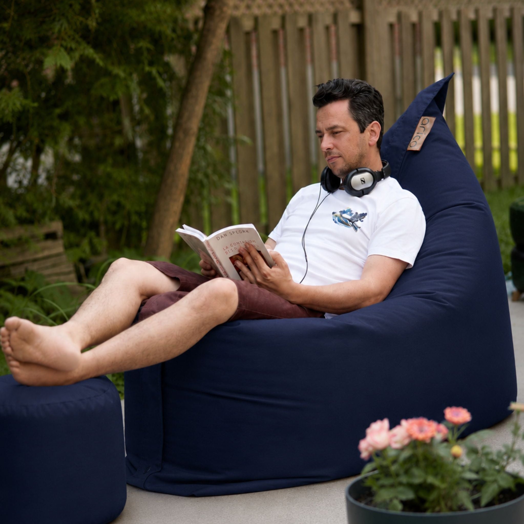 Homme assis lisant un livre confortablement dans un bean bag et un pouf à pieds arico bleu à l'extérieur dans une cours avec des fleurs de jardin rose